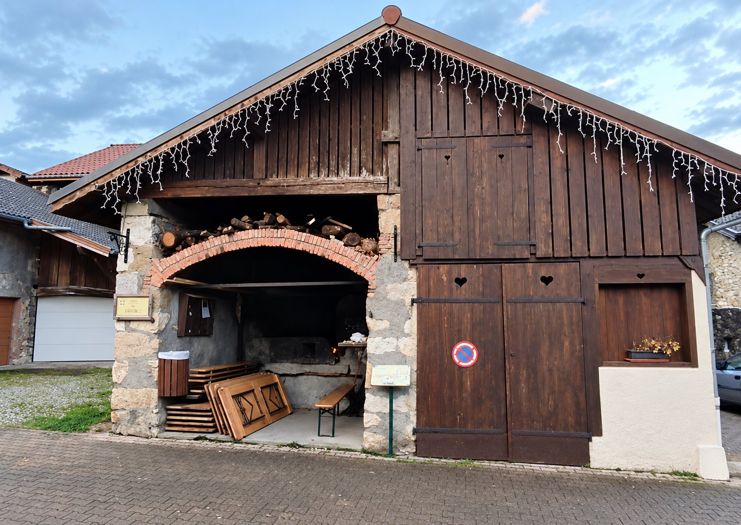 Façade d'une ancienne grange en bois sombre et pierre, avec un porche voûté en brique abritant un banc, des tables pliantes et des bûches, décorée de guirlandes lumineuses.