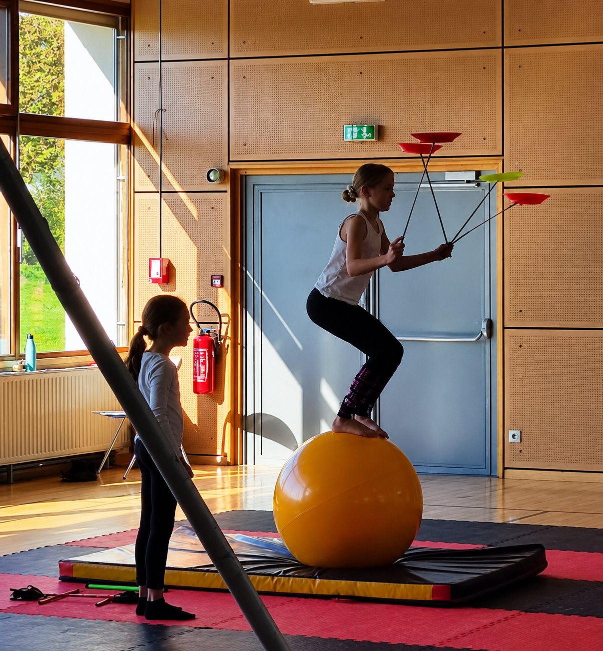 Dans une salle de sport lumineuse, une jeune artiste tient en équilibre sur un gros ballon jaune en faisant tourner des assiettes, pendant qu'une fillette l'observe.
