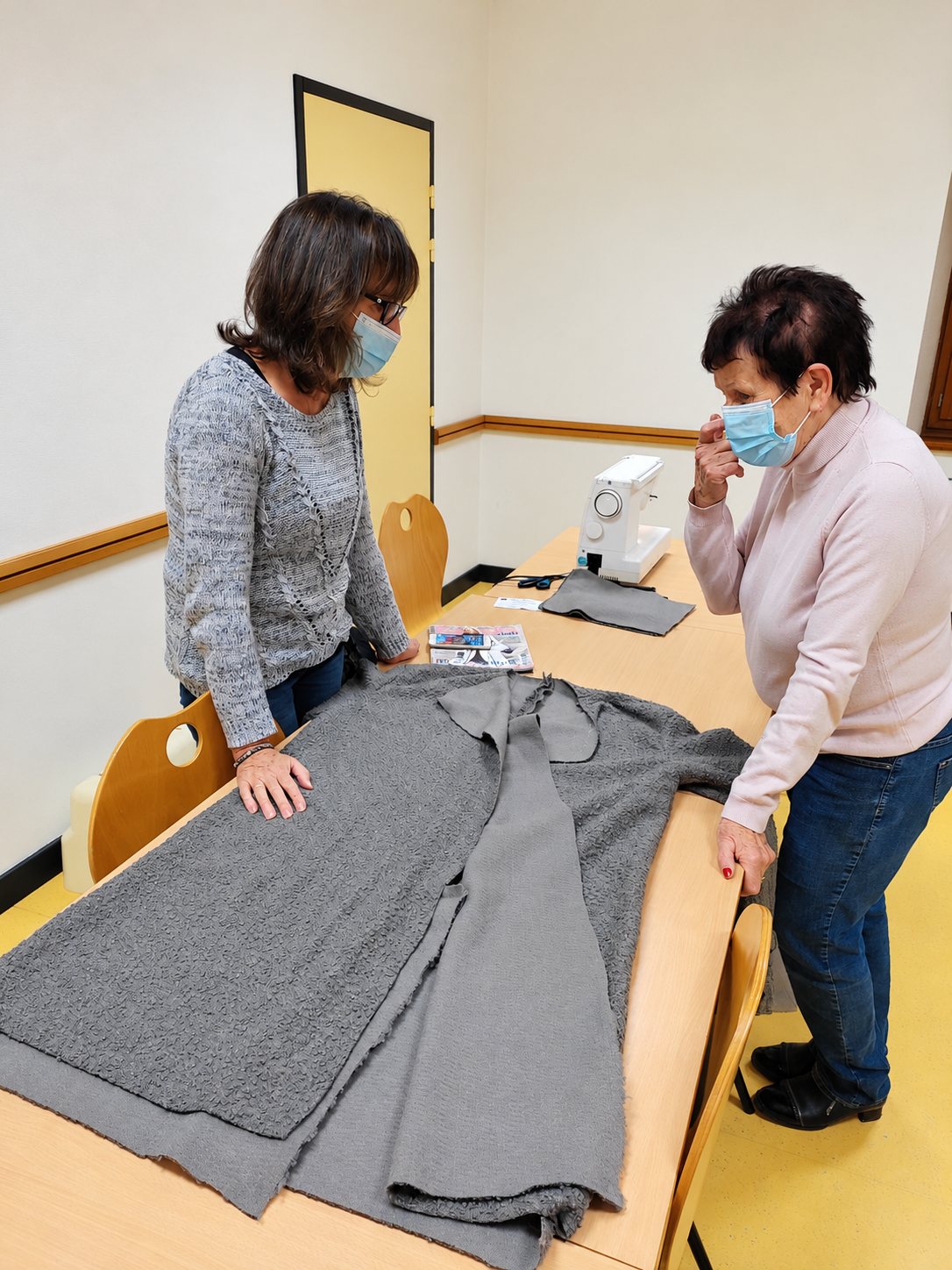 Deux femmes portant un masque observent un grand tissu en laine bouclée grise étalé sur une table, avec une machine à coudre au fond de la salle.