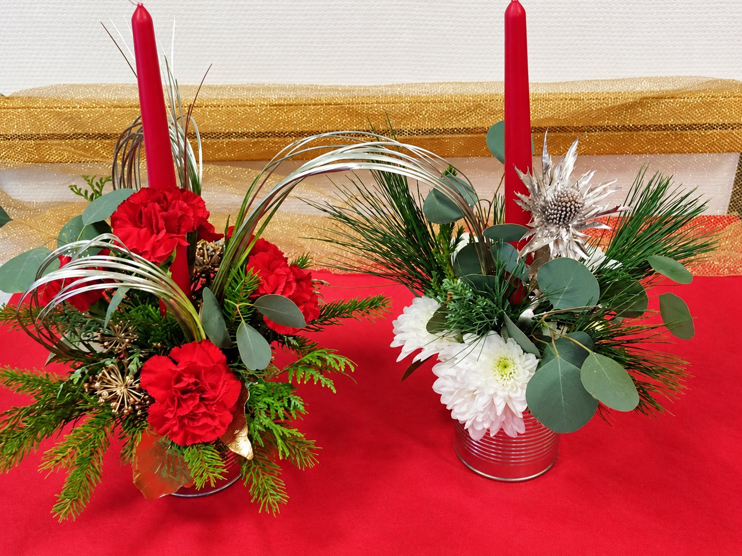 Deux compositions florales de Noël avec bougies rouges, œillets rouges, chrysanthèmes blancs, eucalyptus et branches de sapin posées sur une nappe rouge.