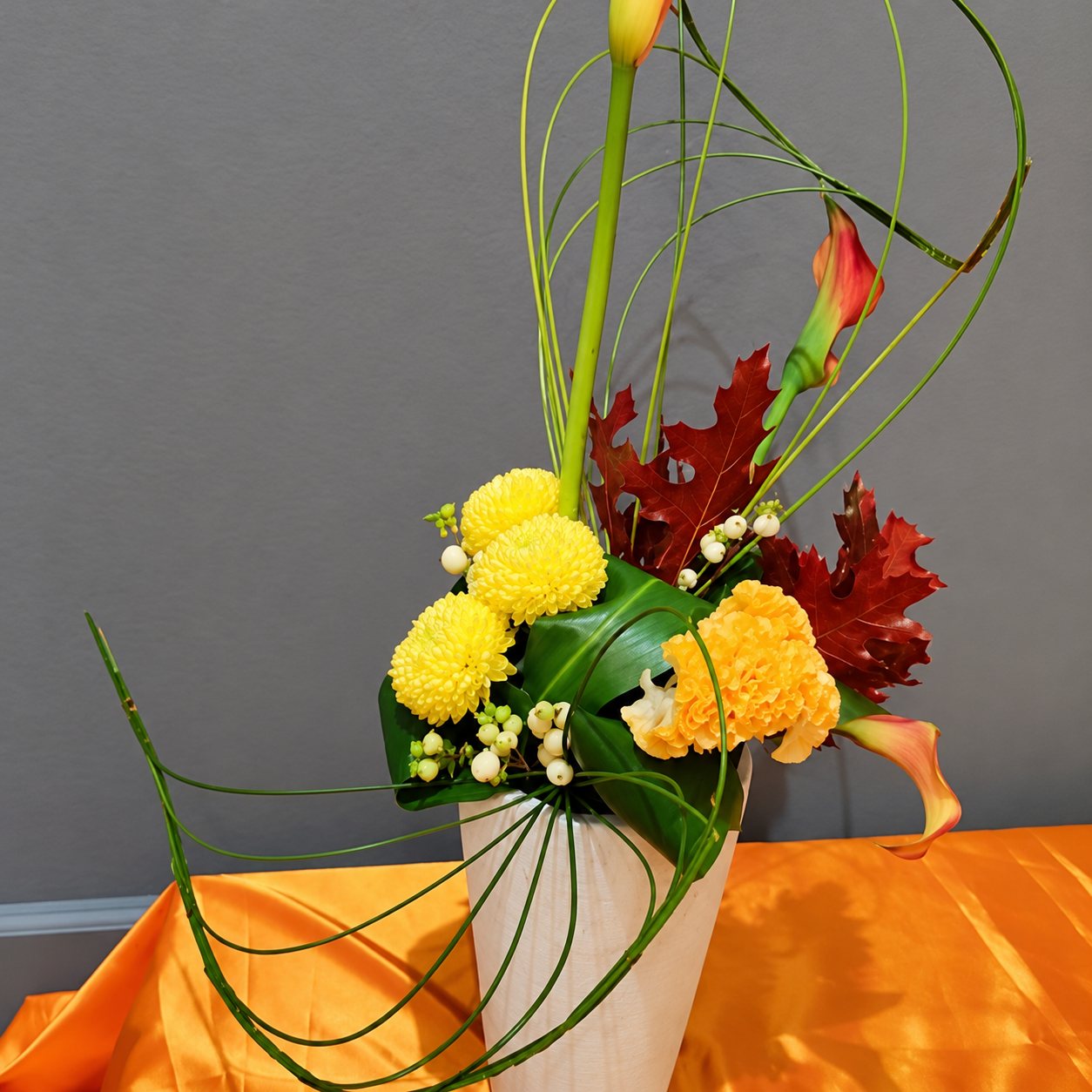 Un arrangement floral aux chrysanthèmes jaunes, feuilles de chêne rouges et arums orangés, présenté dans un vase clair posé sur une table drapée de satin orange.