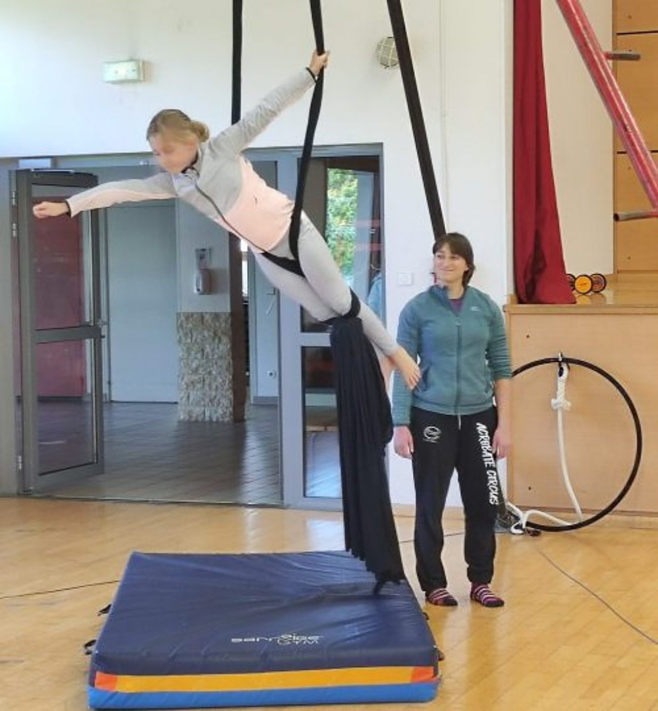 Une jeune fille s'exerce à l'acrobatie aérienne sur un tissu noir au-dessus d'un tapis de réception, sous le regard d'une monitrice en veste bleue dans un gymnase.