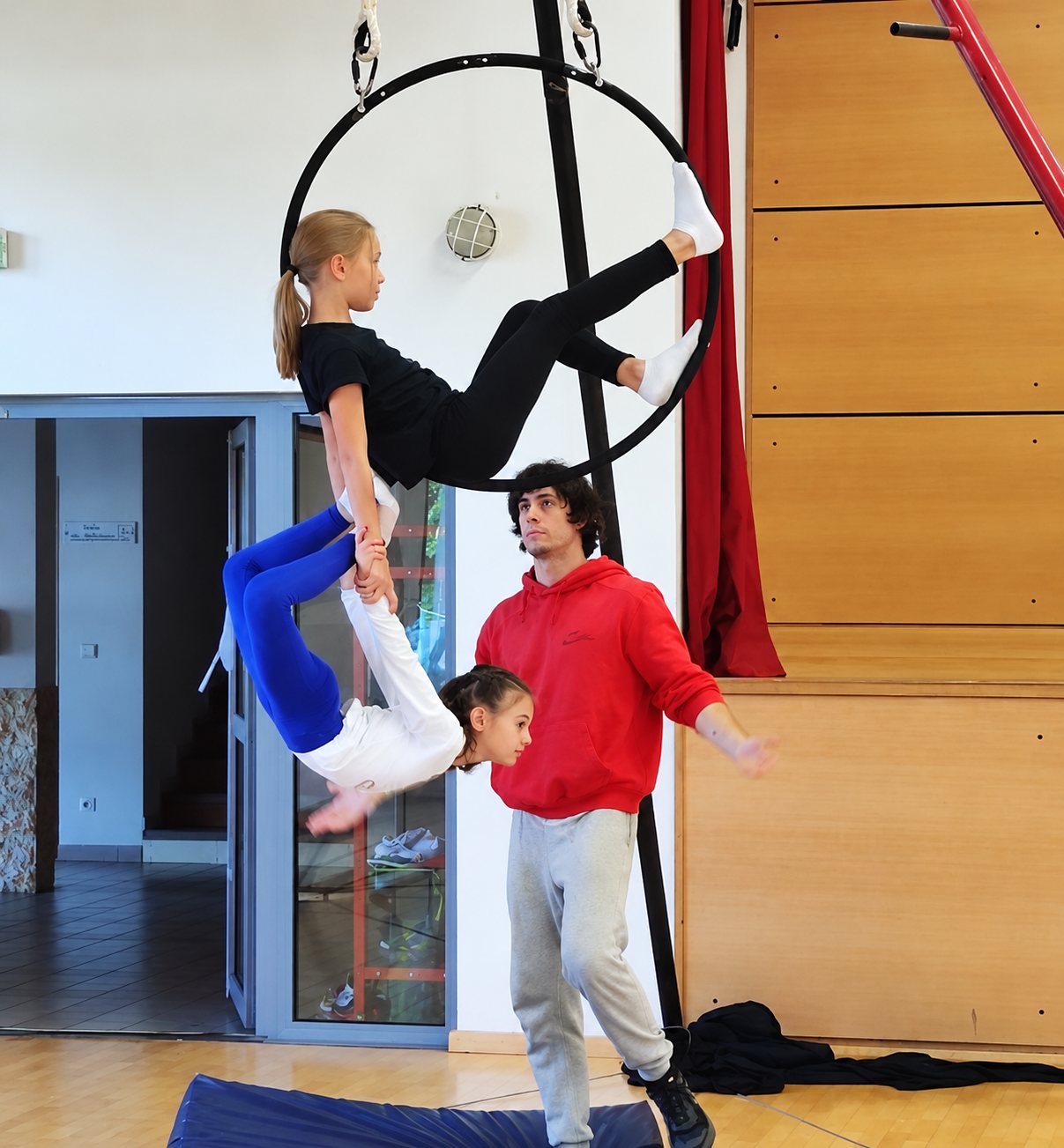 Deux enfants font une figure acrobatique dans un cerceau aérien suspendu, tandis qu'un jeune homme en sweat rouge les surveille dans un gymnase.
