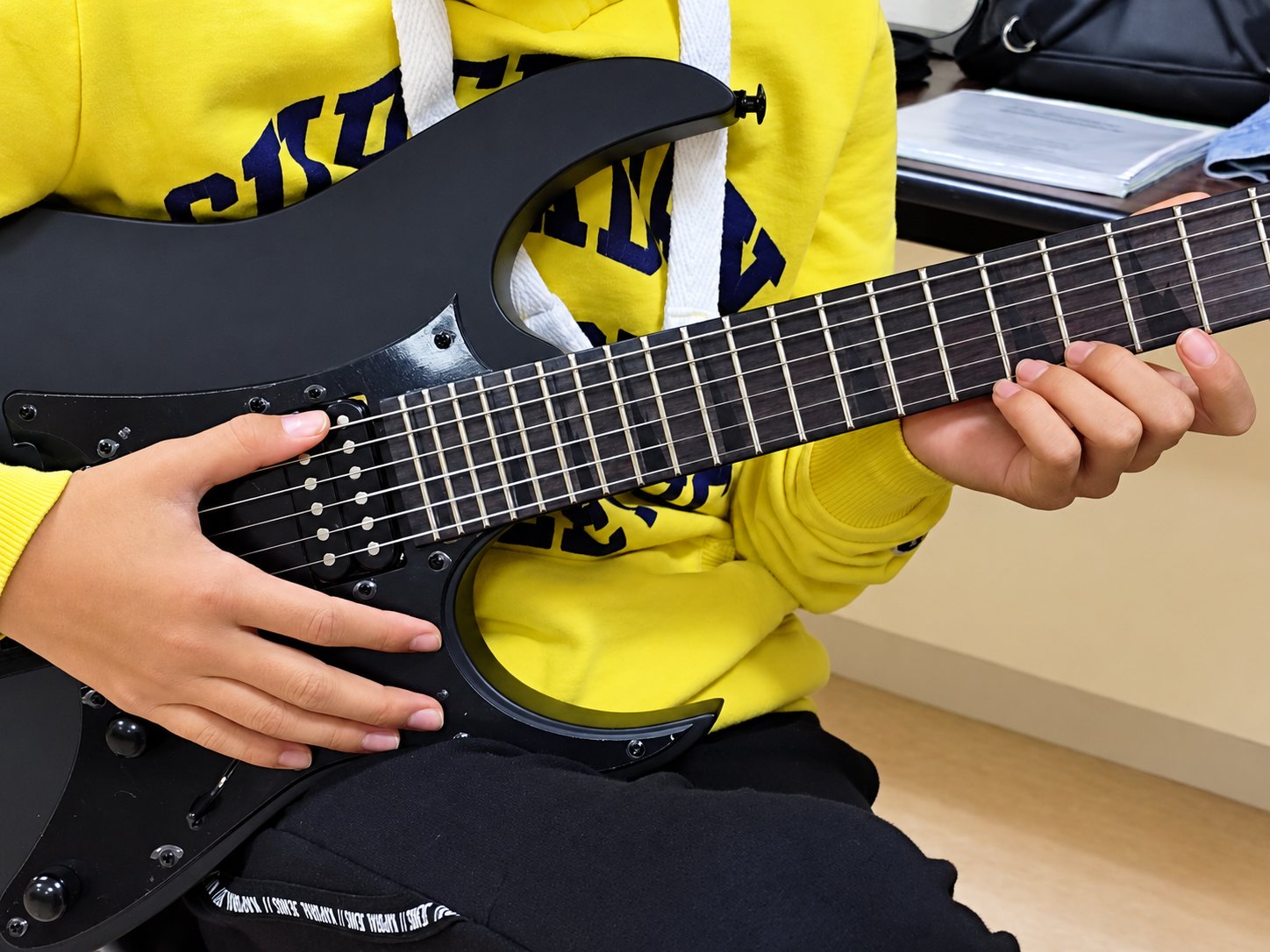 Gros plan sur les mains d'une personne en sweat jaune qui joue d'une guitare électrique noire dans une salle de cours.