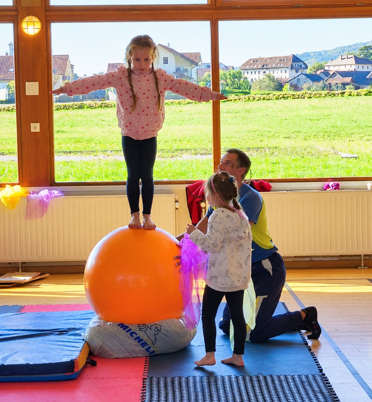 Dans une salle lumineuse aux grandes fenêtres, une fillette se tient debout en équilibre sur un grand ballon orange pendant qu'un adulte et une autre enfant l'observent.