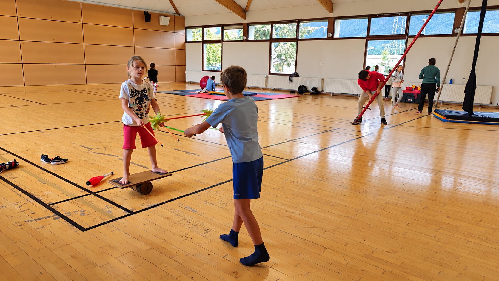 Dans un gymnase, un enfant se tient en équilibre sur une planche à rouleau avec des massues de jonglage pendant qu'un autre enfant lui fait face, et d'autres participants s'exercent au fond de la salle.