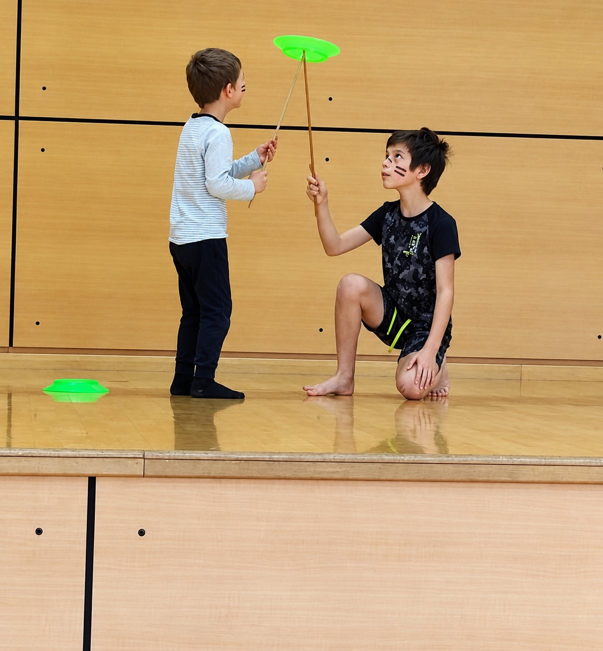 Sur une scène en bois, un garçon à genoux tient une baguette avec une assiette verte en rotation face à un enfant debout.