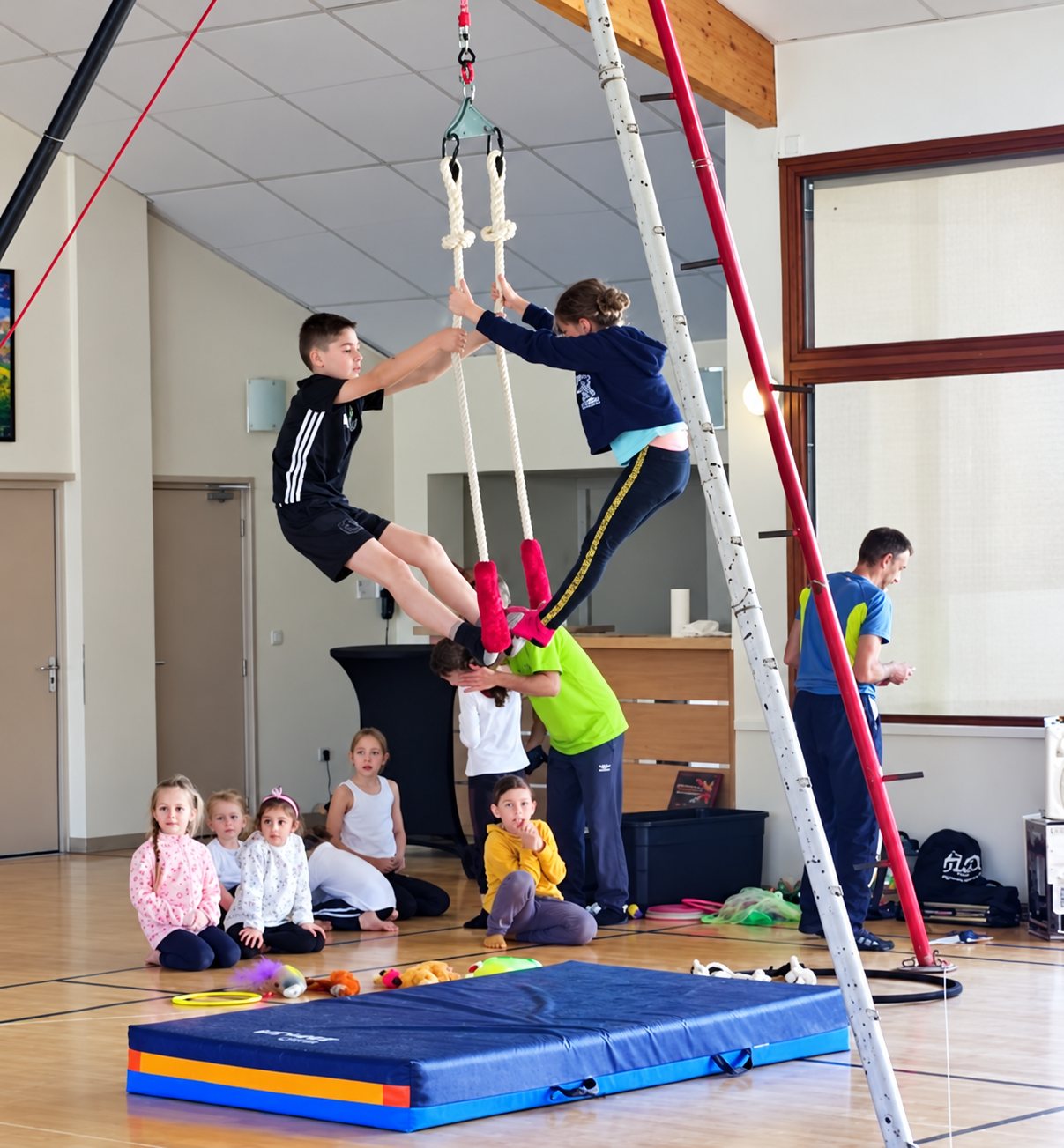 Dans une salle de sport, deux enfants font du trapèze suspendu au-dessus d'un tapis bleu et orange, sous le regard d'un animateur et d'autres enfants assis au sol.