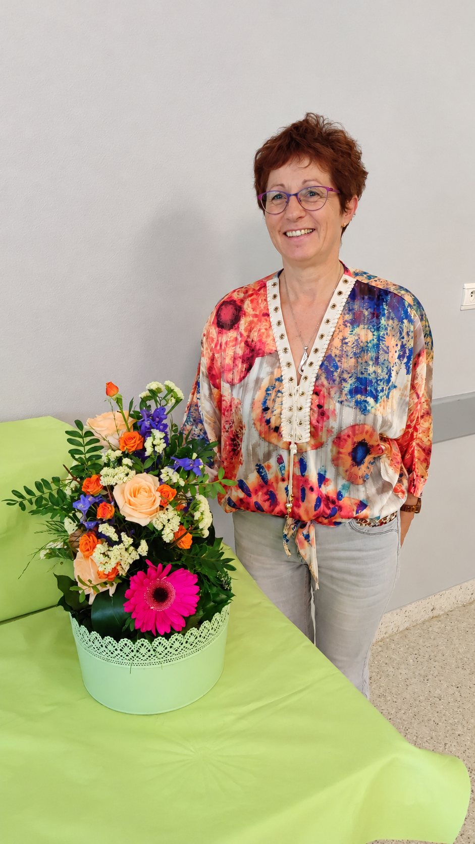 Une femme souriante aux cheveux courts et aux lunettes se tient debout derrière un bouquet de roses, de gerbera rose et de feuillage, posé dans un cache-pot blanc sur une table verte.