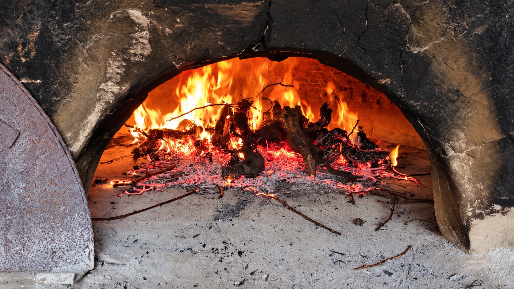Des flammes et des braises brûlent dans un four à bois traditionnel en pierre, dont la porte en métal rouillé est posée à gauche.