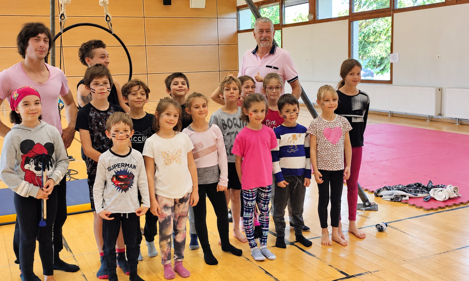 Un groupe d'enfants au visage maquillé pose avec deux adultes dans un gymnase, près d'un cerceau aérien et d'un tapis rouge.