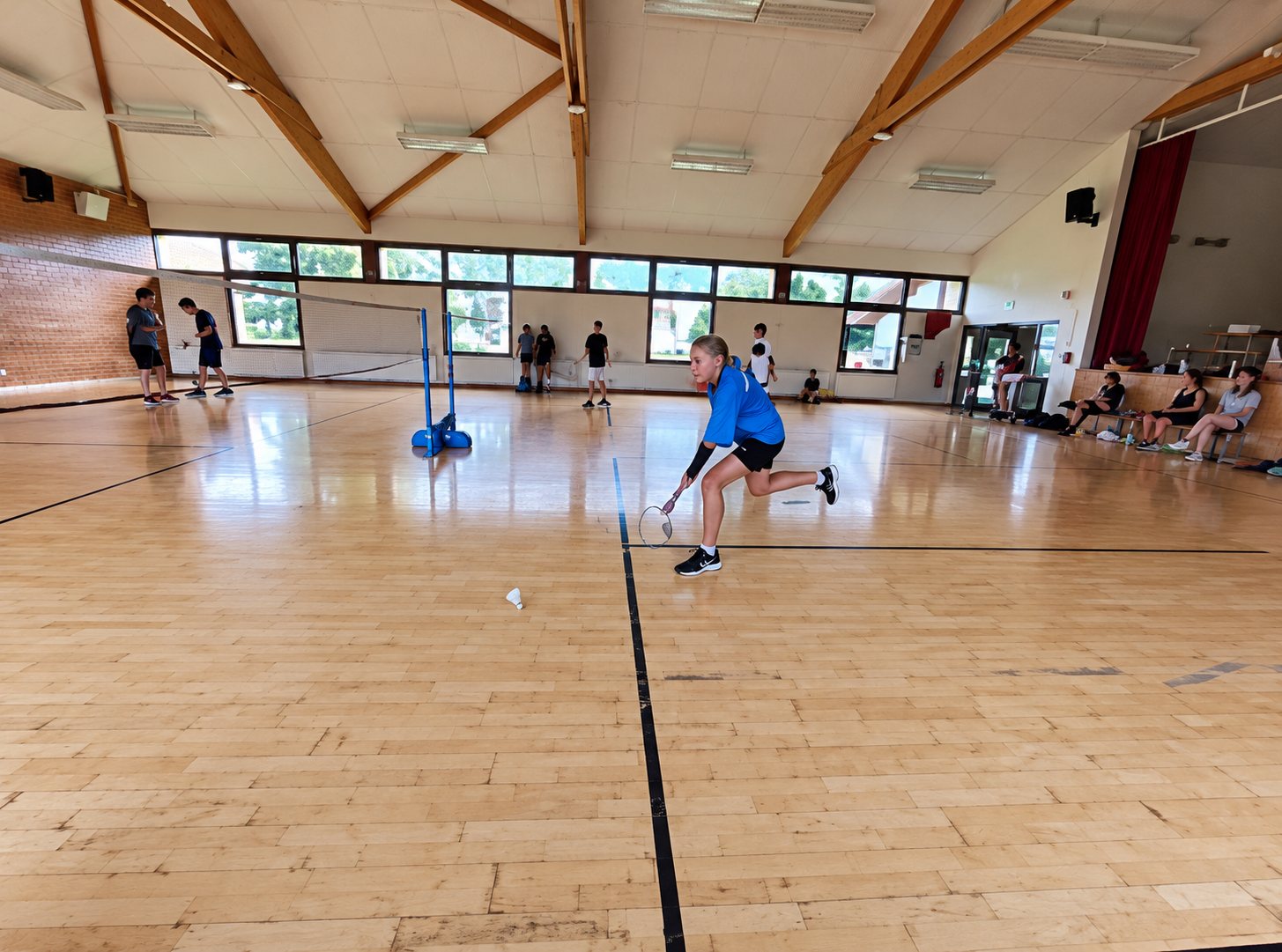 Un joueur en t-shirt bleu court sur le parquet d'un gymnase pendant un match de badminton, avec des spectateurs au fond.