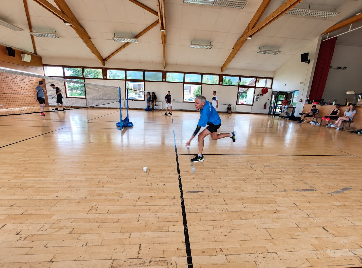 Un joueur en maillot bleu se penche pour récupérer un volant sur le parquet d'un gymnase, avec d'autres joueurs et des spectateurs en arrière-plan.