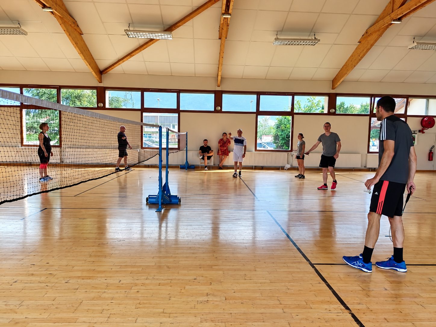 Plusieurs personnes jouent au badminton dans un gymnase lumineux au parquet en bois, autour d'un filet bleu.