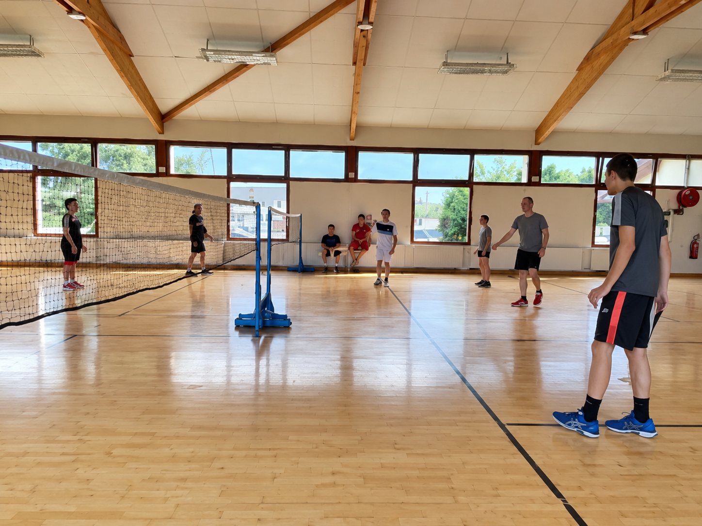 Plusieurs personnes jouent au badminton dans une salle de sport lumineuse au parquet clair et à la charpente en bois, avec un filet au centre.