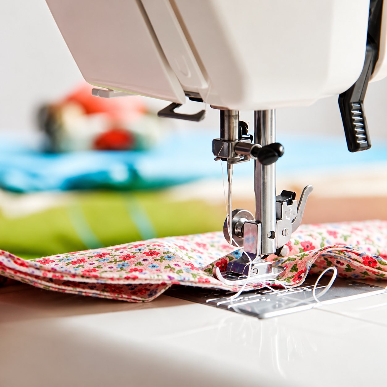 Gros plan sur le pied-de-biche d'une machine à coudre blanche en train de piquer un tissu à petites fleurs roses, avec des tissus colorés flous à l'arrière-plan.