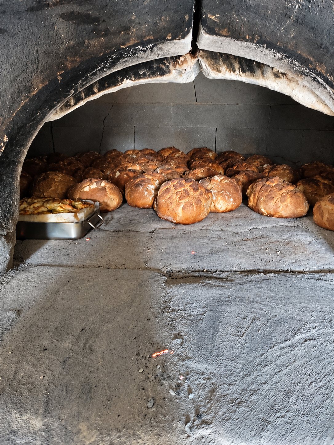 Plusieurs pains ronds dorés cuisent au fond d'un four à bois en pierre, à côté d'un plat métallique.