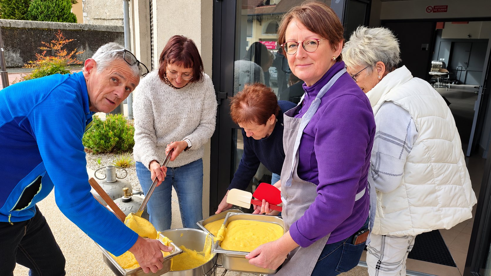 Cinq personnes se relayent à l'extérieur pour remuer et répartir une préparation jaune, de la polenta, dans des bacs en inox posés près d'une bouteille de gaz.
