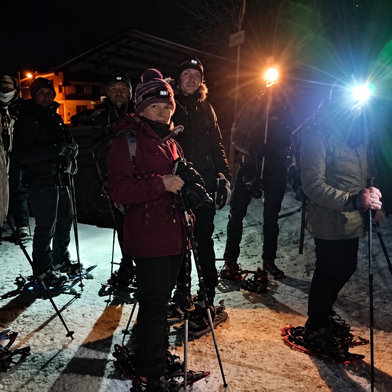 Un groupe de personnes chaudement vêtues, munies de bâtons et de lampes frontales, se tient sur la neige en pleine nuit avant une sortie en raquettes.