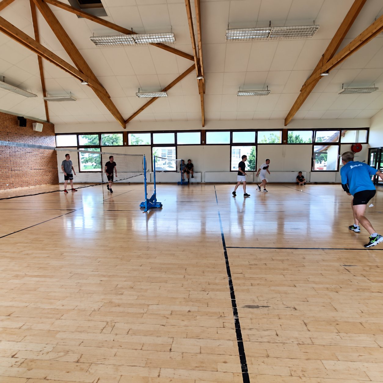 Des personnes jouent au badminton dans une grande salle de sport au parquet clair, sous une charpente en bois apparente.